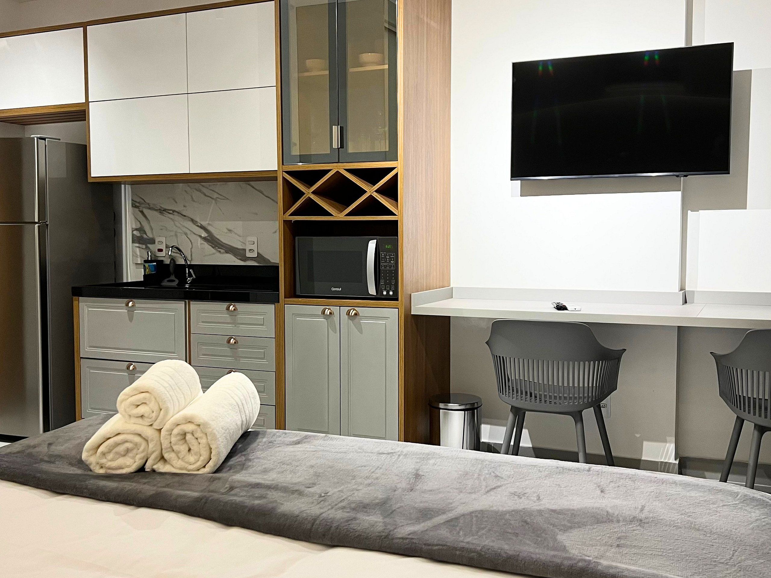 A modern hotel room with a bed, a flat-screen TV, a kitchenette, and a seating area.