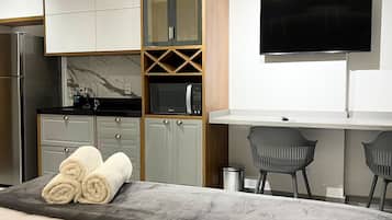 A modern hotel room with a bed, a flat-screen TV, a kitchenette, and a seating area.