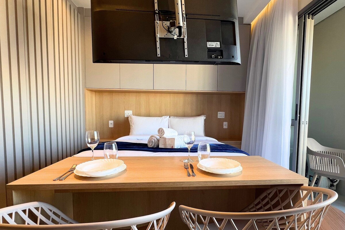 Una habitación de hotel con cama, mesita de noche con vasos, una silla y un televisor sobre la cama.