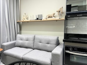 A modern living room with a grey sofa, a small table, and a kitchen area with a microwave and oven. - GRU Residence by Housi (Guarulhos)