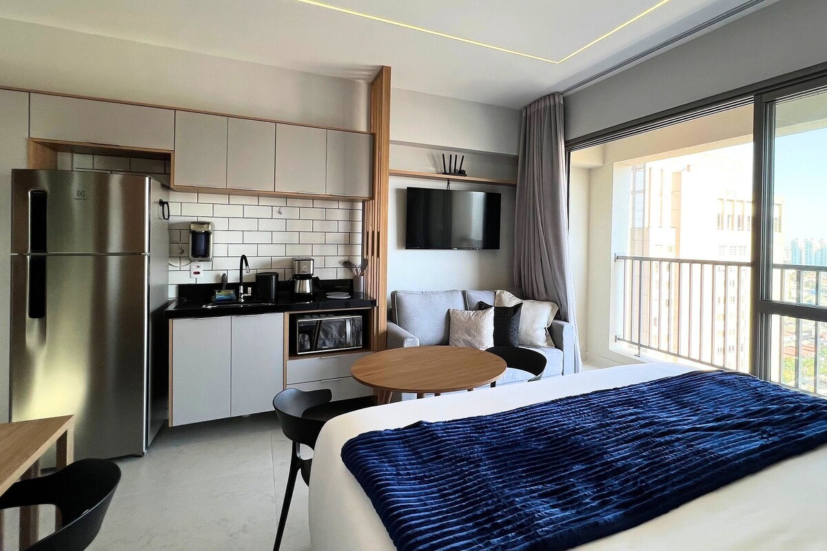 Una habitación de hotel moderna con kitchenette, cama, zona de estar y un ventanal grande.