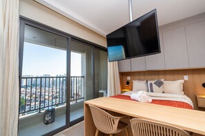 A hotel room with a large window, a bed with a red and white bedspread, a wooden desk, two chairs, a television, and a city view. - GRU Residence by Housi (Guarulhos)