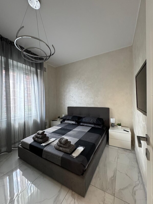 A modern bedroom with a large bed, a bedside table, a wall-mounted TV, and a ceiling light fixture. - HBPeri Apartments (Torino)