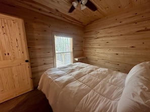 A room with wooden walls, a bed with a white duvet, a ceiling fan, and a window with blinds. - Two Bedroom Cabin - Cozy cabin equipped with most necessities, sleeps 6 - with a view! (Andalusia)