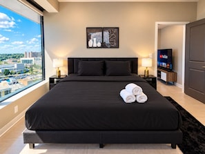 A modern bedroom with a large bed, two bedside tables, a TV, and a view of the city. - Downtown Afterglow | 2 Queen 2BR (Birmingham)
