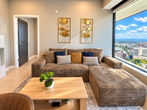 A modern living room with a sectional sofa, a coffee table, and a view of a cityscape through large windows. - The City Perch | Queen + Queen 2BR (Birmingham)