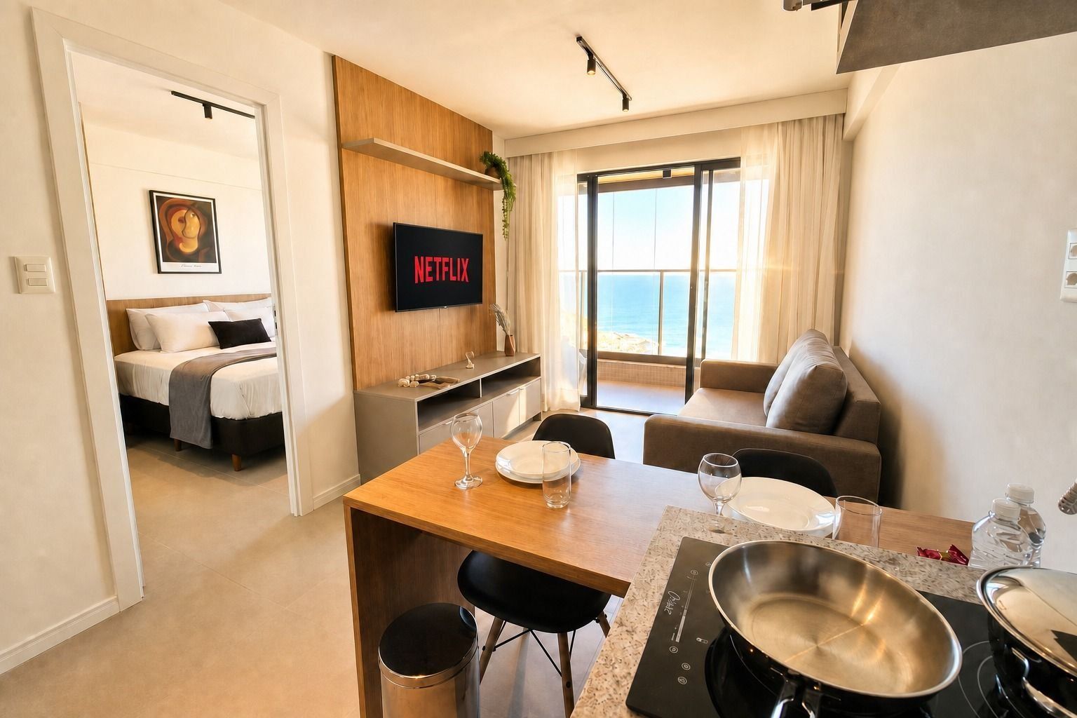 Quarto de hotel moderno com cama, televisão, área de refeições com mesa e cadeiras, e vista para o mar pela janela.
