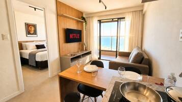Quarto de hotel moderno com cama, televisão, área de refeições com mesa e cadeiras, e vista para o mar pela janela.