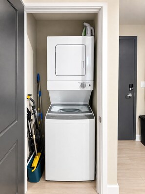 A compact laundry room with a front-loading washer and dryer, a mop, and a broom. - Magic City Highlife | King Bed + 3BR (Birmingham)