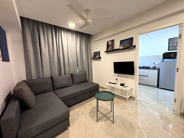 A modern living room with a sectional sofa, a small round table, a flat-screen TV, and a ceiling fan. - Finland 1BHK_Hiranandani Estate_City View (Thane)