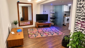 A living room with a flat-screen TV on a wooden stand, a patterned rug, a wooden table with a laptop, and a mirror reflecting the room.