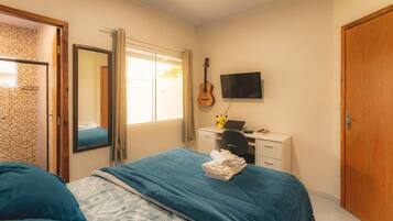 Quarto com cama, escrivaninha com computador, guitarra e televisão na parede.