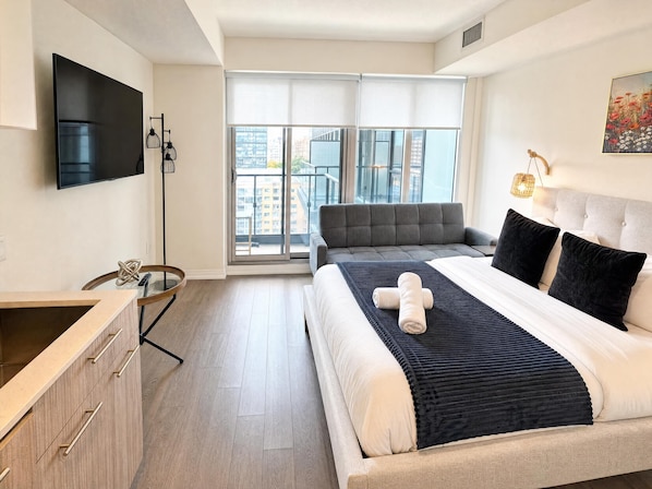 A modern bedroom with a large bed, a flat-screen TV, a sofa, and a balcony with a city view. - StayToronto - Downtown Jarvis Place (Toronto)