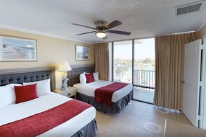 Quarto de hotel com duas camas, ventilador de teto, mesa de cabeceira com abajur e uma janela grande com vista.