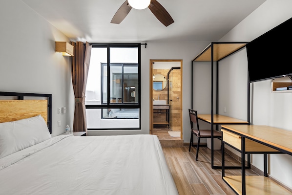 Habitación de hotel con cama, escritorio, silla, ventilador de techo, ventana y baño visible a través de la puerta.