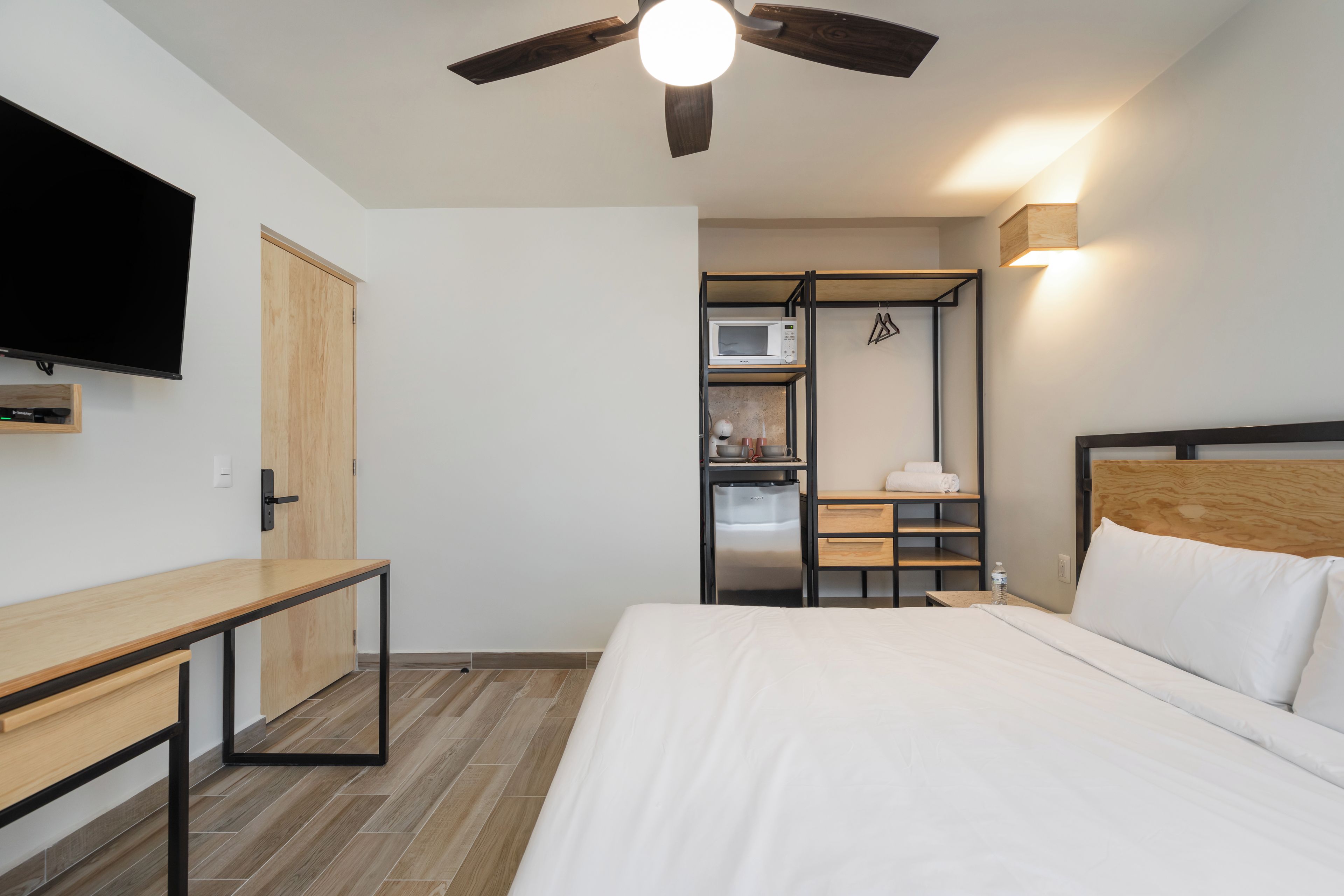 Un dormitorio con cama, escritorio, televisor, ventilador de techo, microondas y refrigerador pequeño.