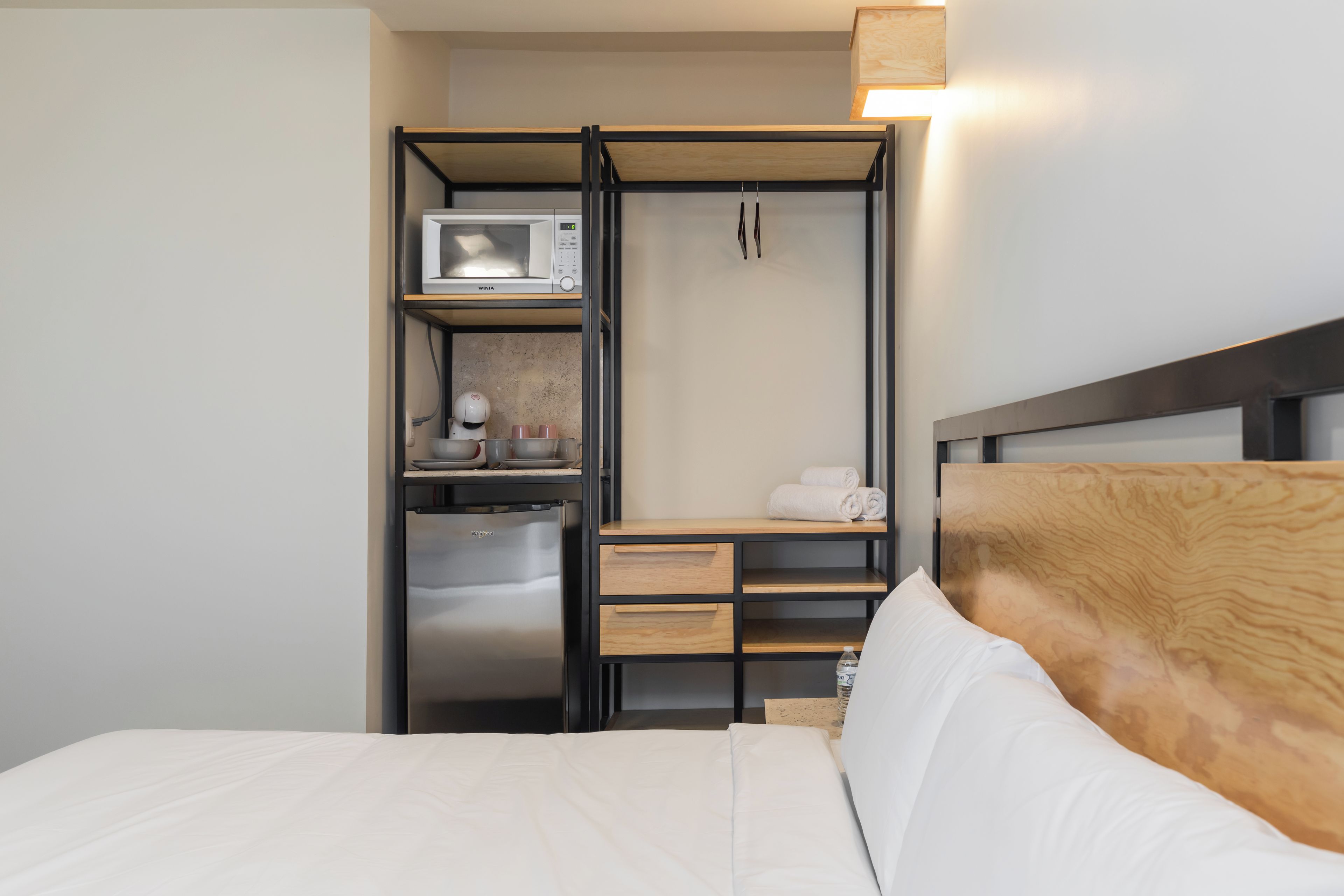 Quarto de hotel com cama, cozinha compacta com micro-ondas e geladeira, e cabeceira de madeira.