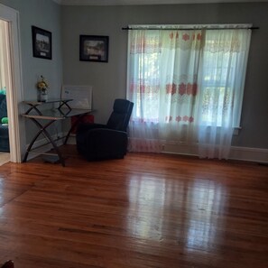 Una habitación con piso de madera, un escritorio con computadora, una silla y dos cuadros en la pared.