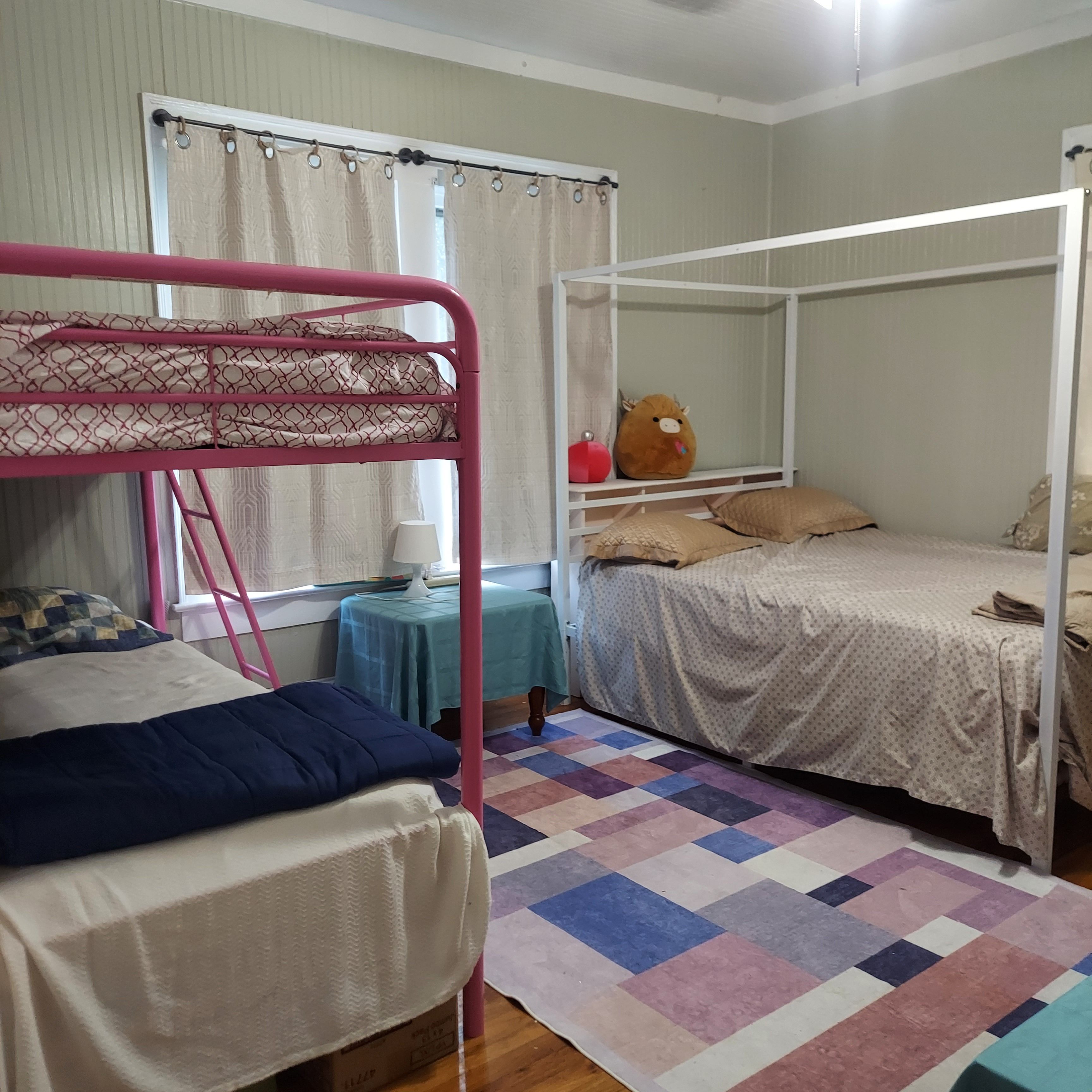Une chambre avec des lits superposés, une petite table avec une lampe, un ours en peluche et un tapis à carreaux.