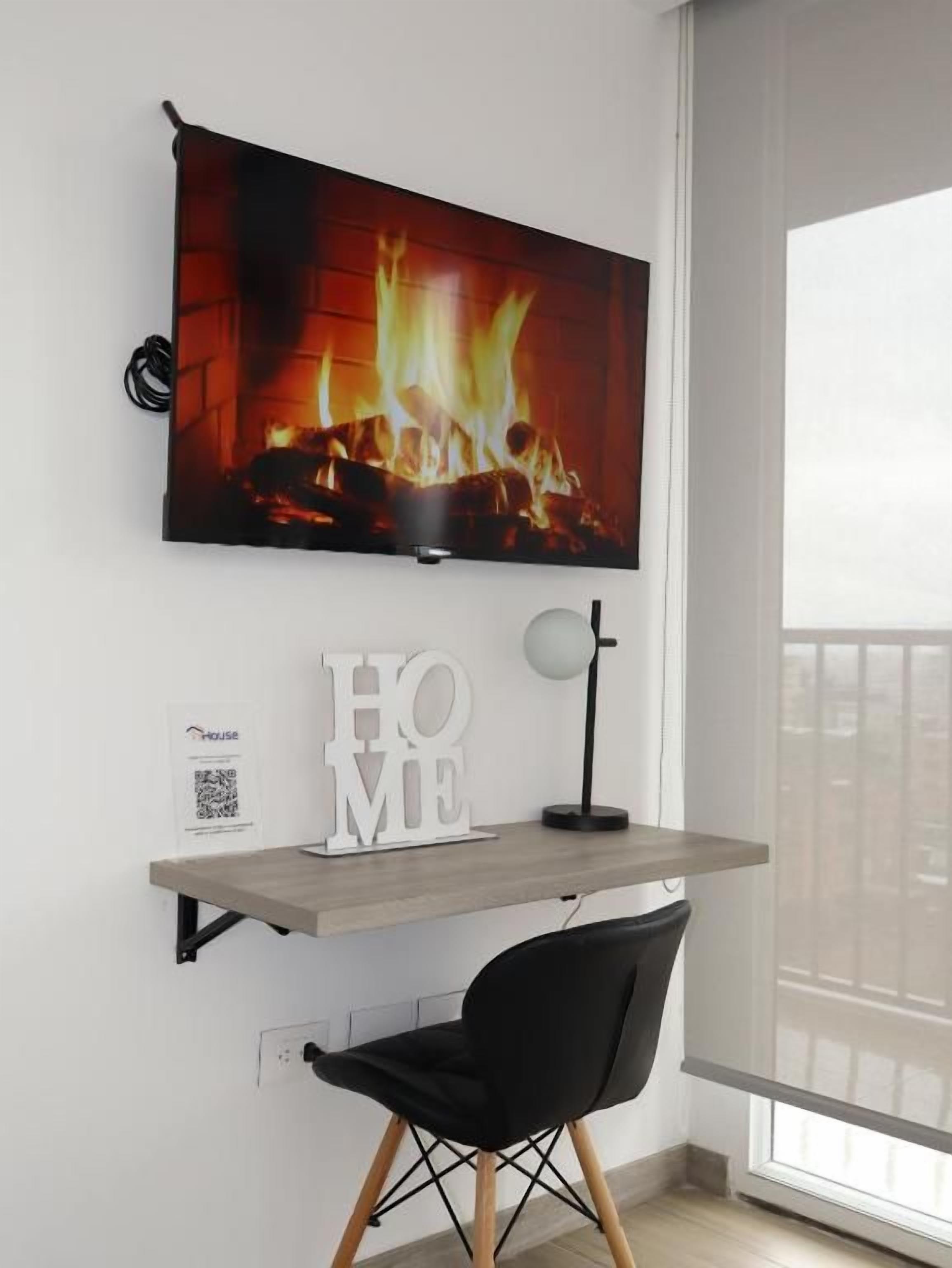 Een moderne kamer met een wandgemonteerde televisie waarop een haard te zien is, een bureau met een stoel en een raam met uitzicht.