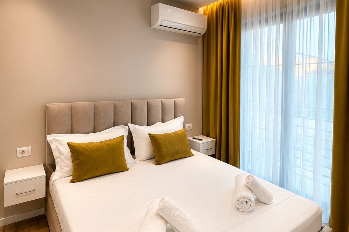 A hotel room with a large bed, a bedside table, an air conditioning unit, and a window with curtains.