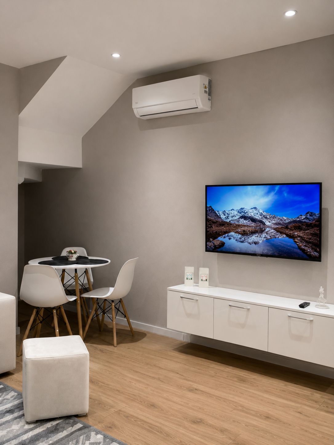 Un saló modern amb un televisor de pantalla plana penjat a la paret, una zona de menjador amb una taula rodona i cadires, i un moble blanc amb calaixos.