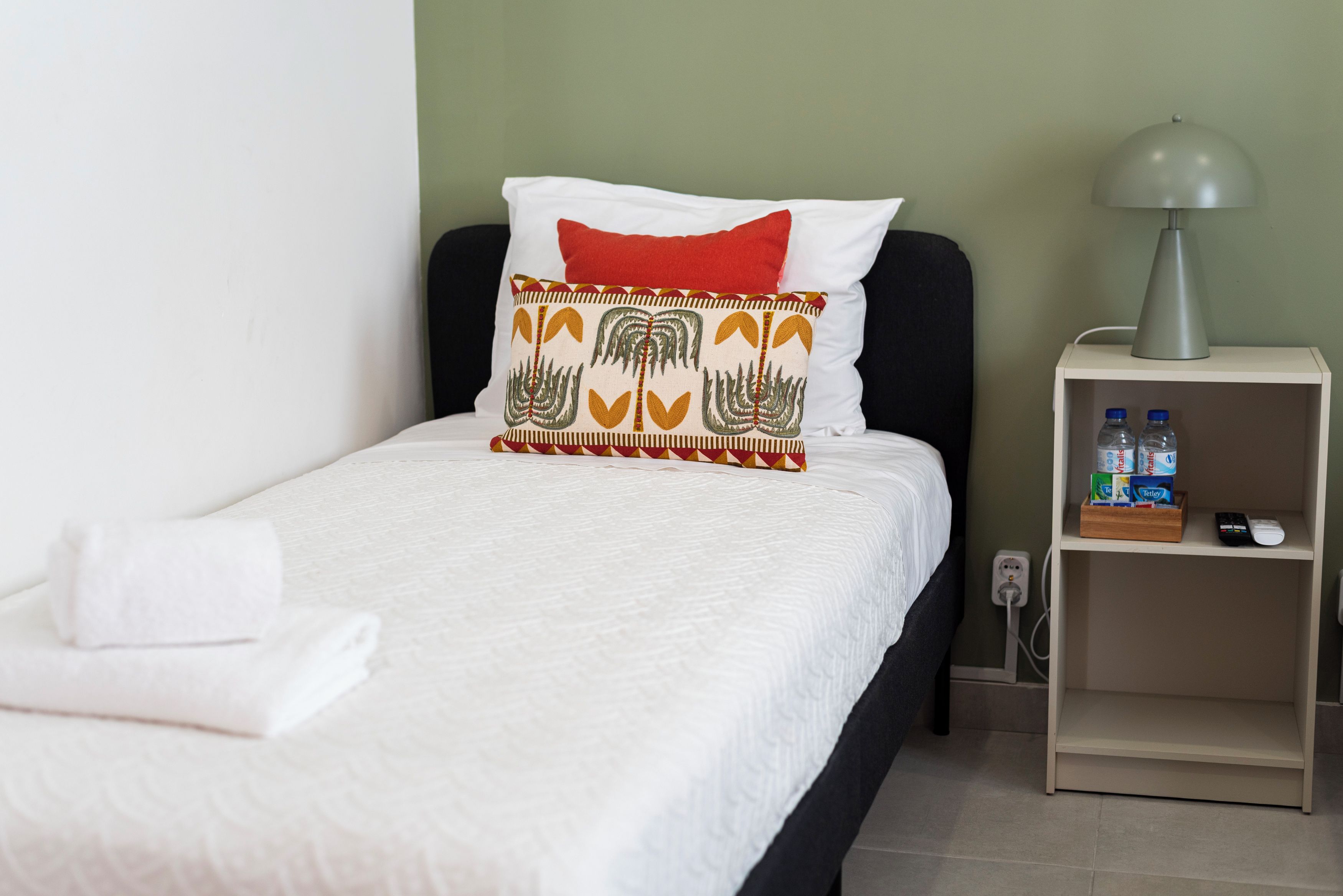 Un lit bien fait, avec une literie blanche et un coussin décoratif, une table de chevet avec une lampe et une bouteille d’eau, et une prise de courant sur le mur.