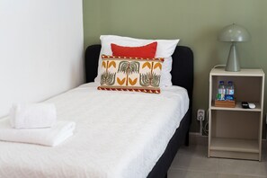 A neatly made bed with white bedding and a decorative pillow, a bedside table with a lamp and bottled water, and a power outlet on the wall.