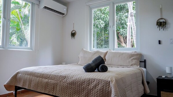 A bedroom with a bed, two windows, a wall-mounted air conditioner, and a small bedside table with a lamp.