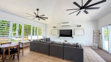 A spacious living room with a large grey sectional sofa, a wooden dining table with chairs, a flat-screen TV mounted on the wall, and two ceiling fans.