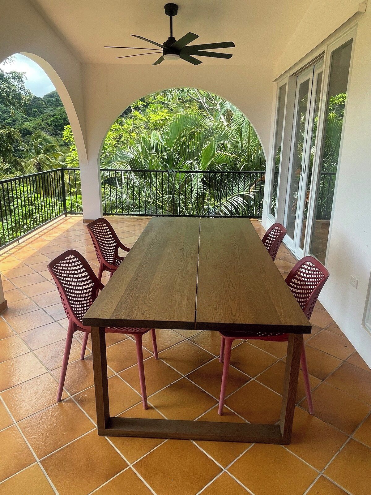 Uma sala de jantar com uma mesa de madeira e cadeiras cor-de-rosa, um ventilador de teto e vista para um verdejante jardim através de um arco.