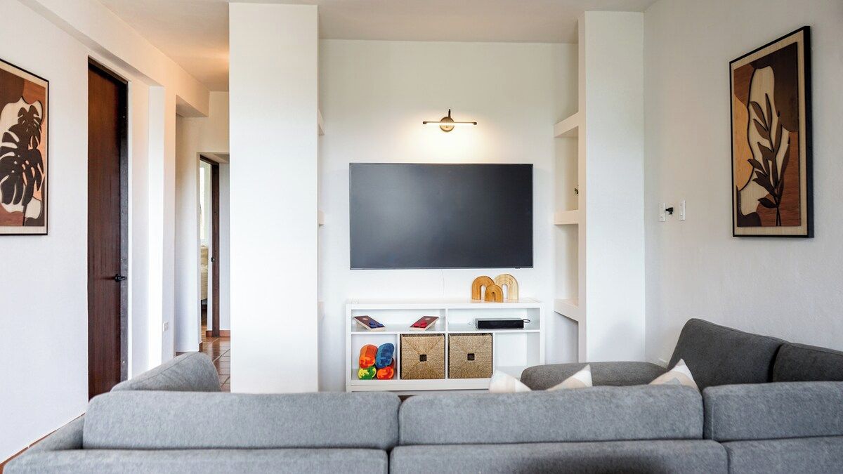 A living room with a grey sofa, a flat-screen TV mounted on a white wall, and a small white entertainment unit below.