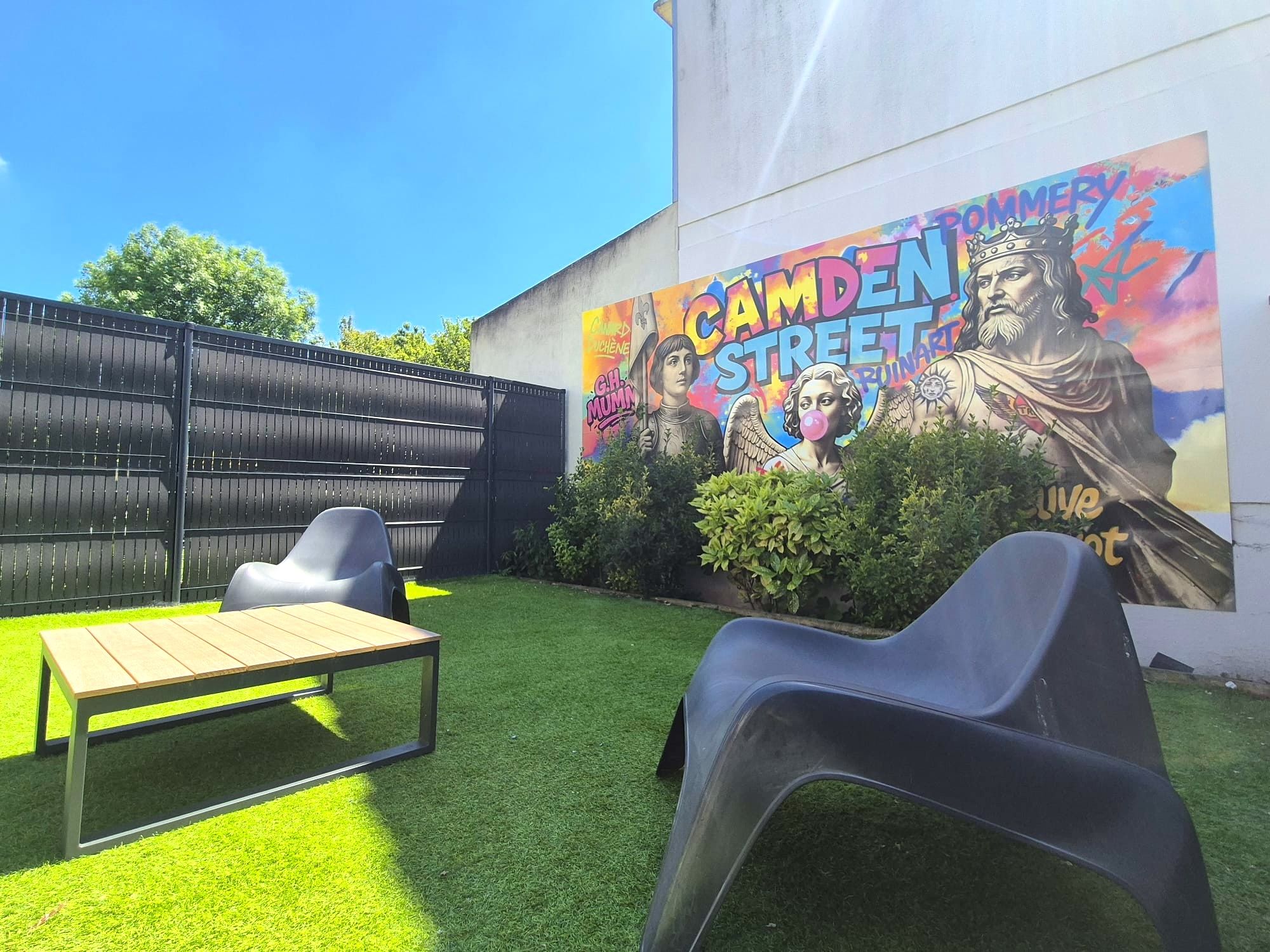 A backyard with artificial grass, a wooden table, two plastic chairs, a black metal fence, and a colorful mural on the wall.