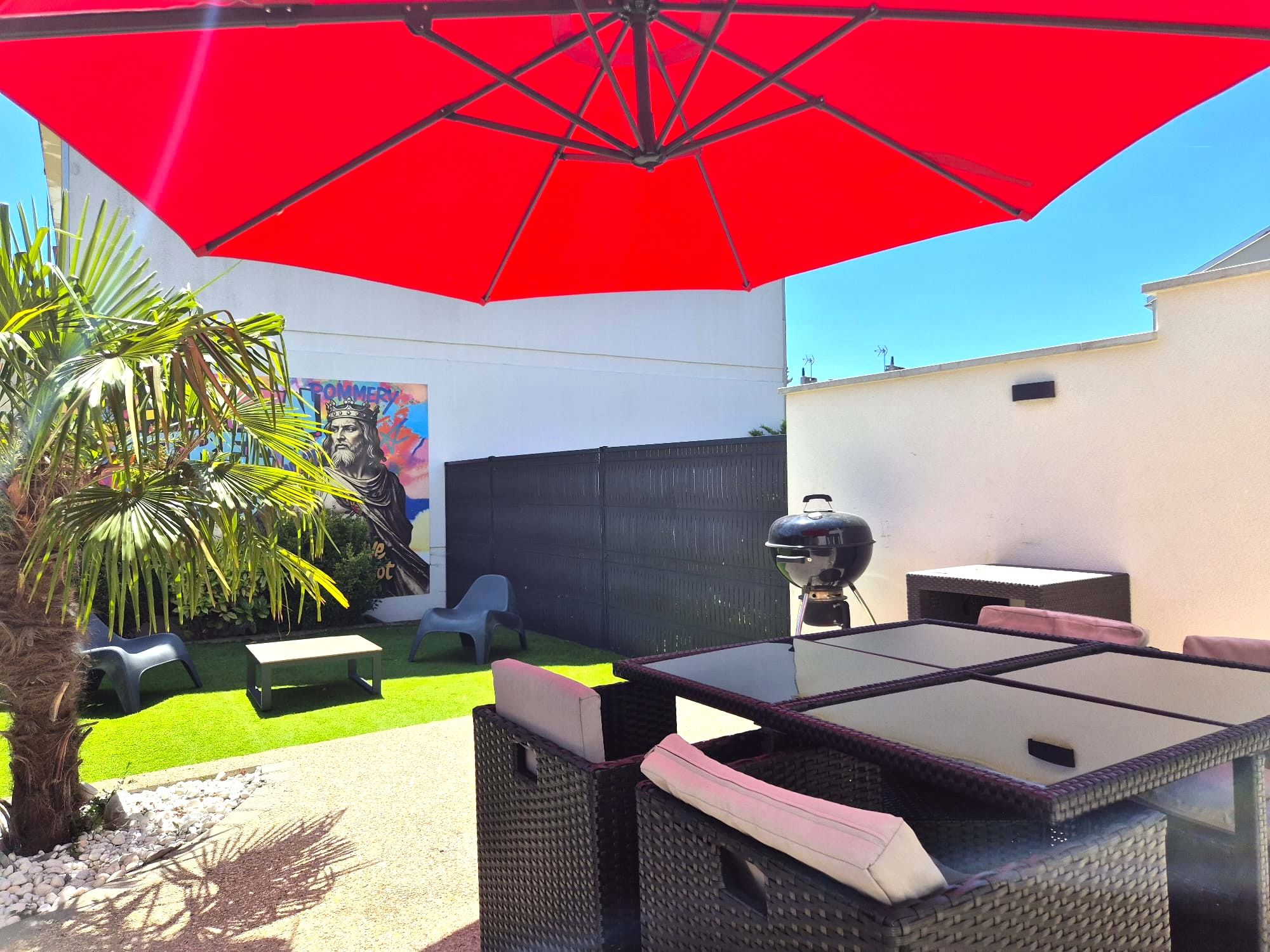 Une terrasse avec une table et des chaises, un parasol rouge, un barbecue et une fresque murale.