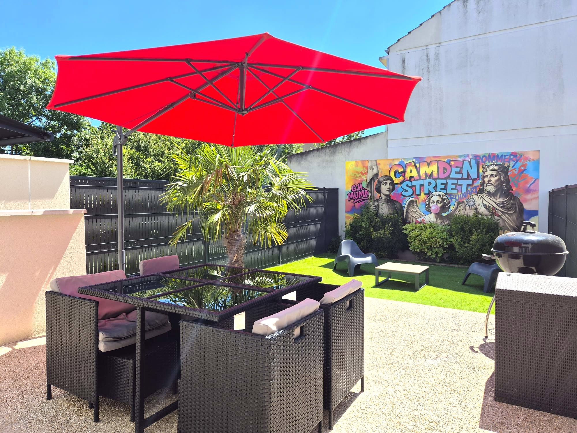 Eine Terrasse im Freien mit Korbmöbeln, einem roten Sonnenschirm und einem Wandgemälde einer Straßenszene.