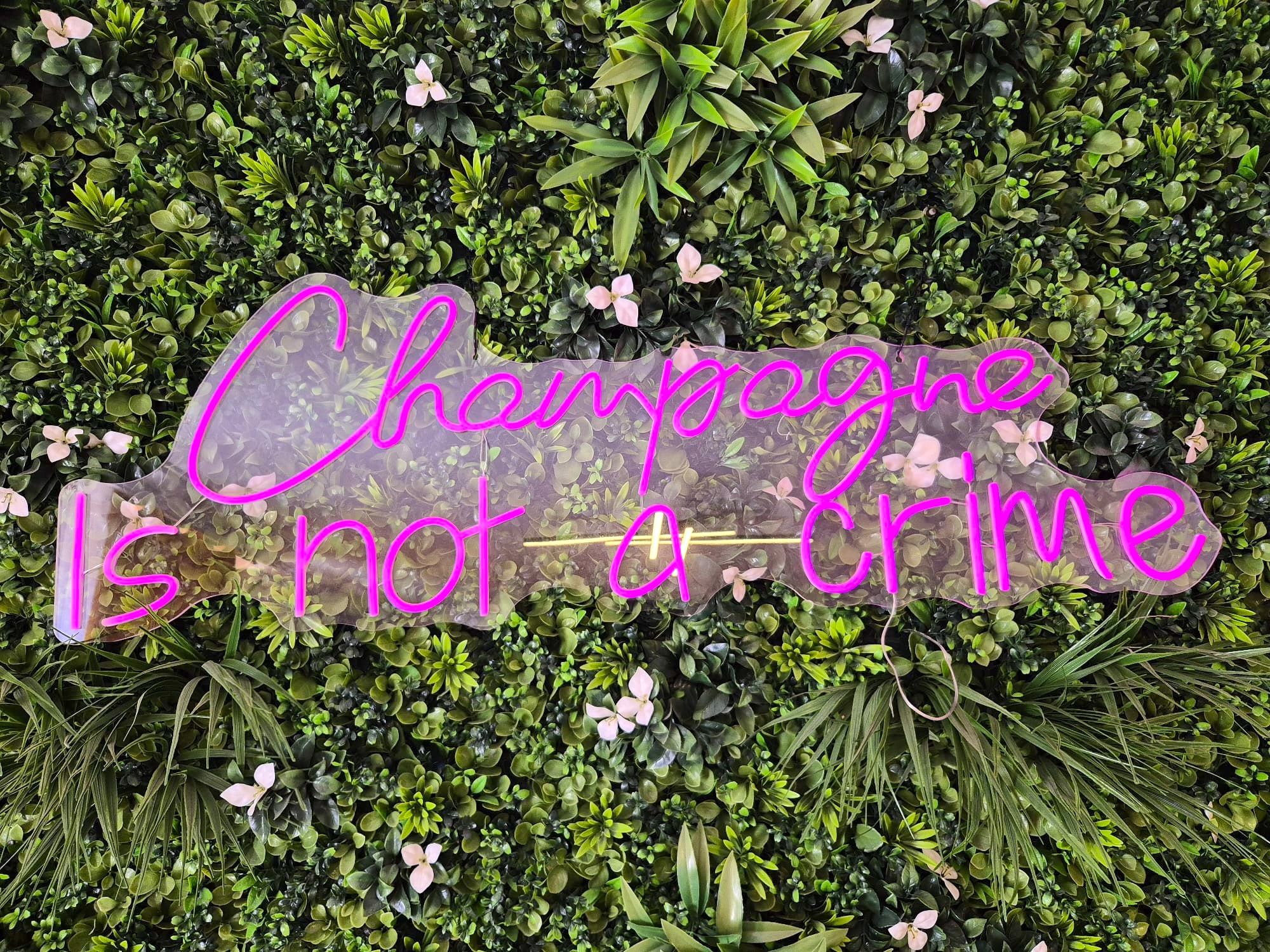 Un letrero de neón con el texto 'Champagne is not a crime' sobre un fondo de follaje verde y pequeñas flores rosadas.