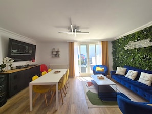 Ein modernes Wohnzimmer mit einem blauen Sofa, einem Esstisch mit gelben Stühlen und einer Küchenzeile, auf deren Fernseher die Serie 'Stranger Things' läuft.