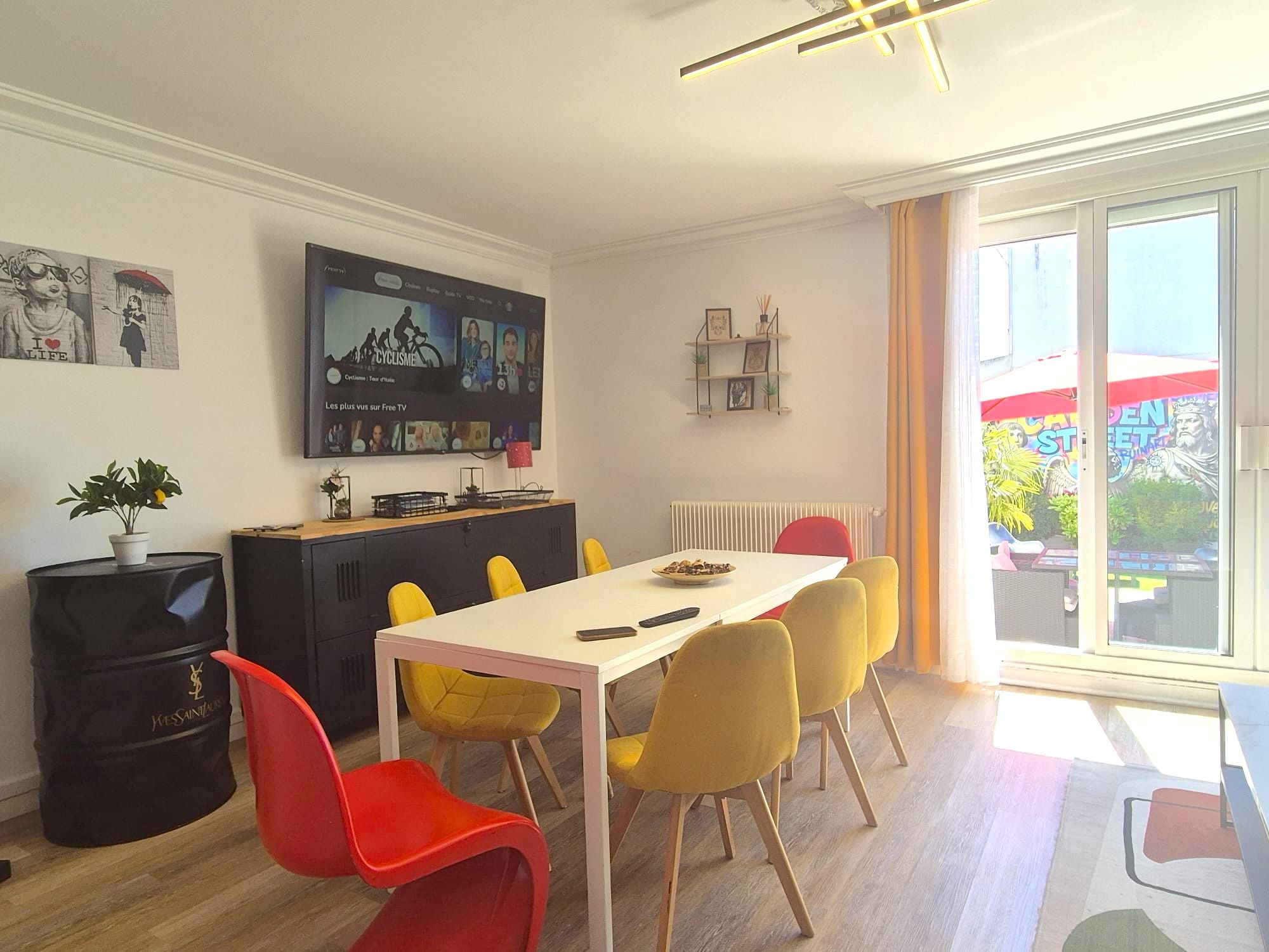 Een moderne woonkamer met een eethoek, een flatscreen televisie aan de muur, een groot raam met uitzicht op buiten, en een zithoek met rode en gele stoelen.
