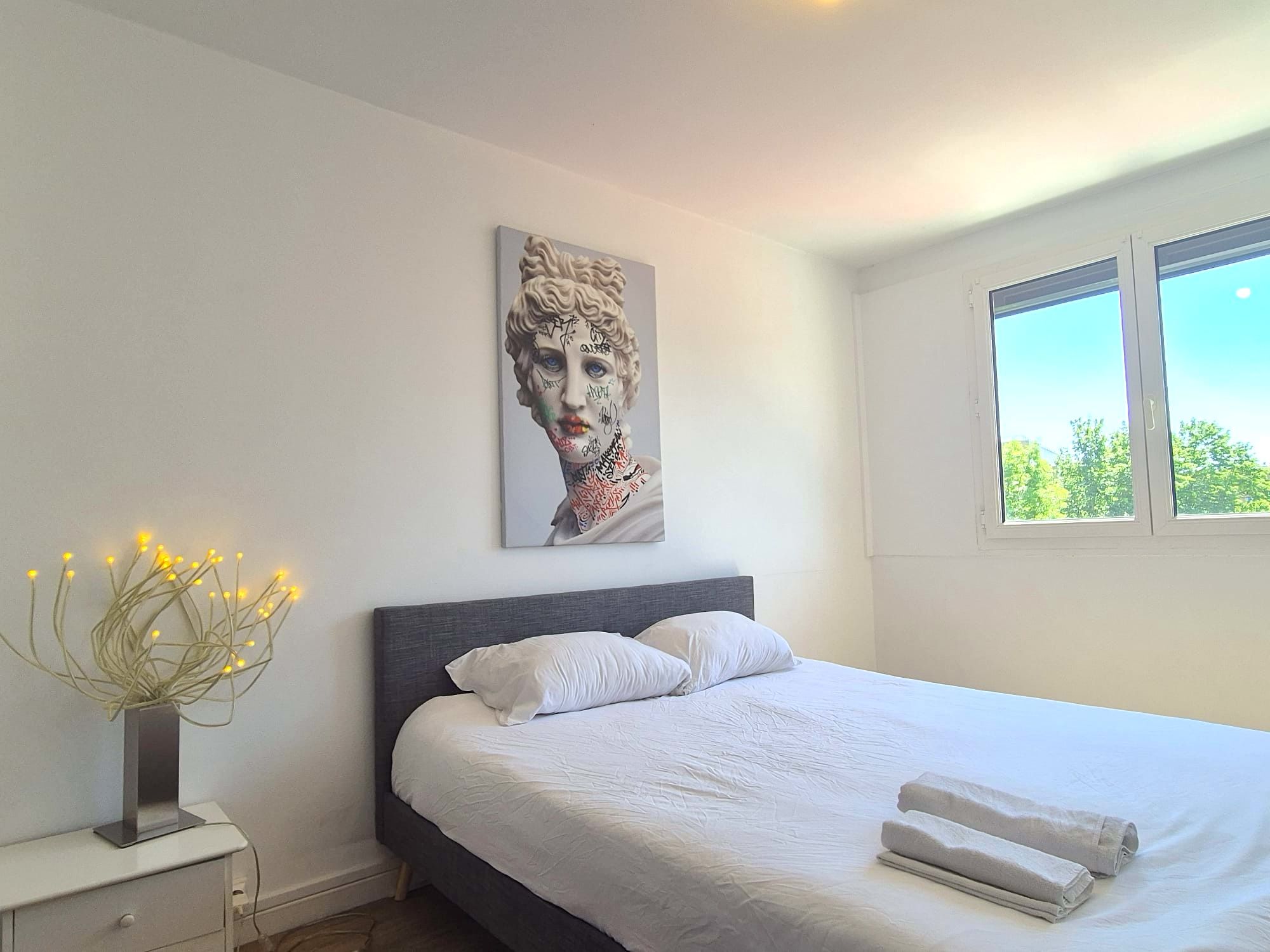 Un dormitorio con cama, almohadas, mesita de noche, un jarrón con luces y un cuadro en la pared.