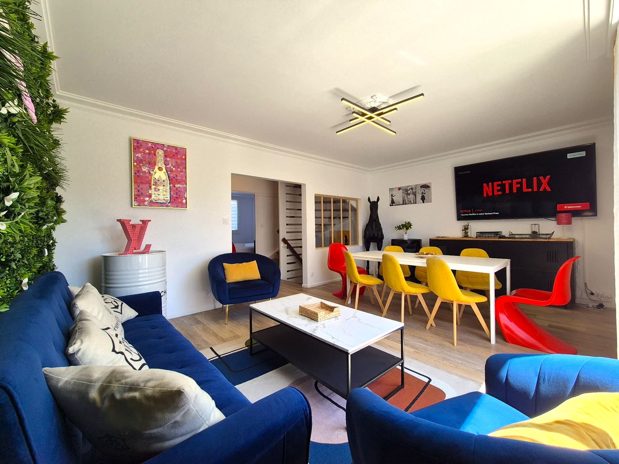 Un salon moderne avec un canapé, une table basse et un coin repas avec des chaises. Un téléviseur affichant Netflix est fixé au mur.