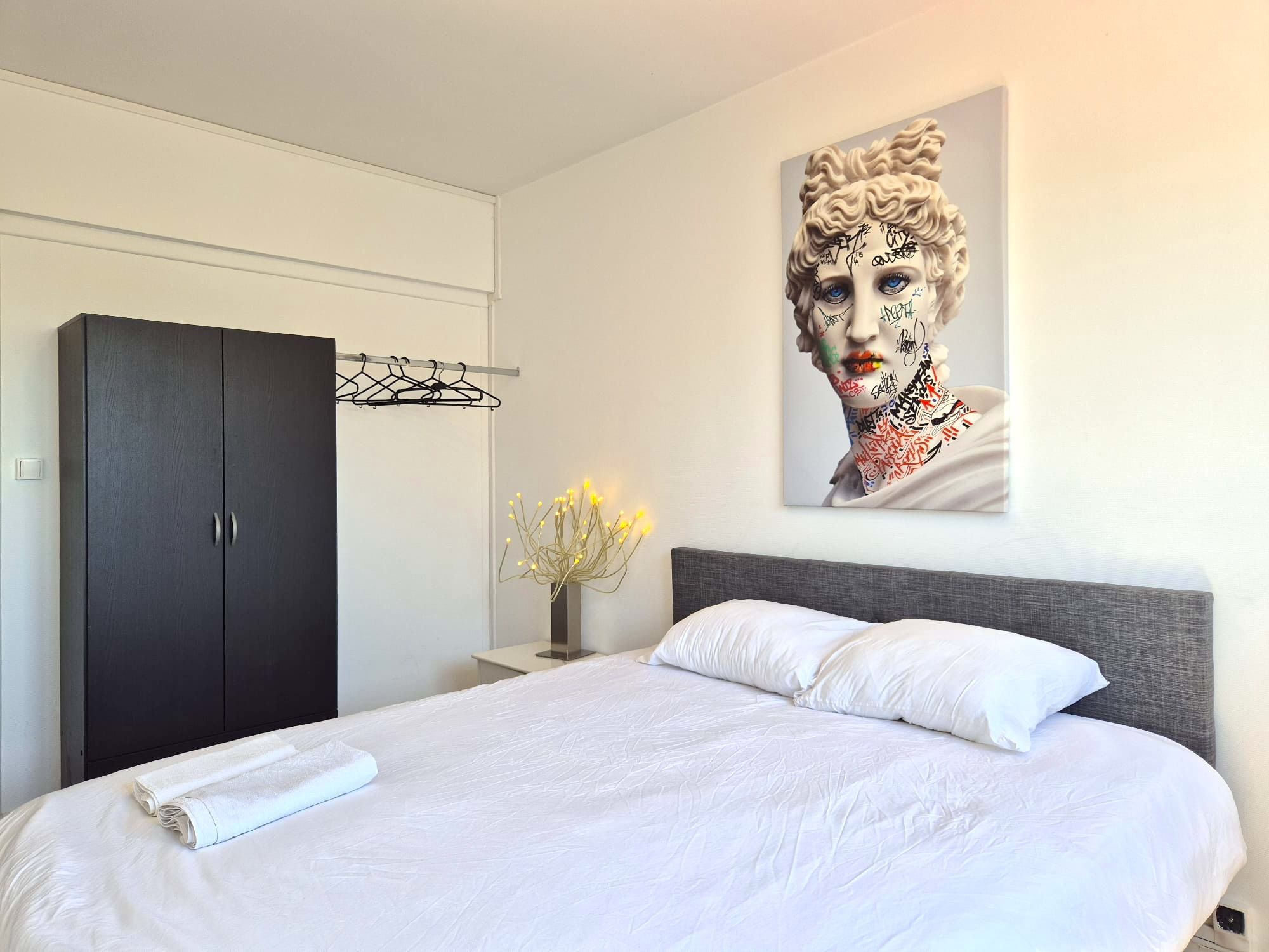 Een slaapkamer met een bed, een garderobe, een nachtkastje met een vaas en een kunstwerk aan de muur.
