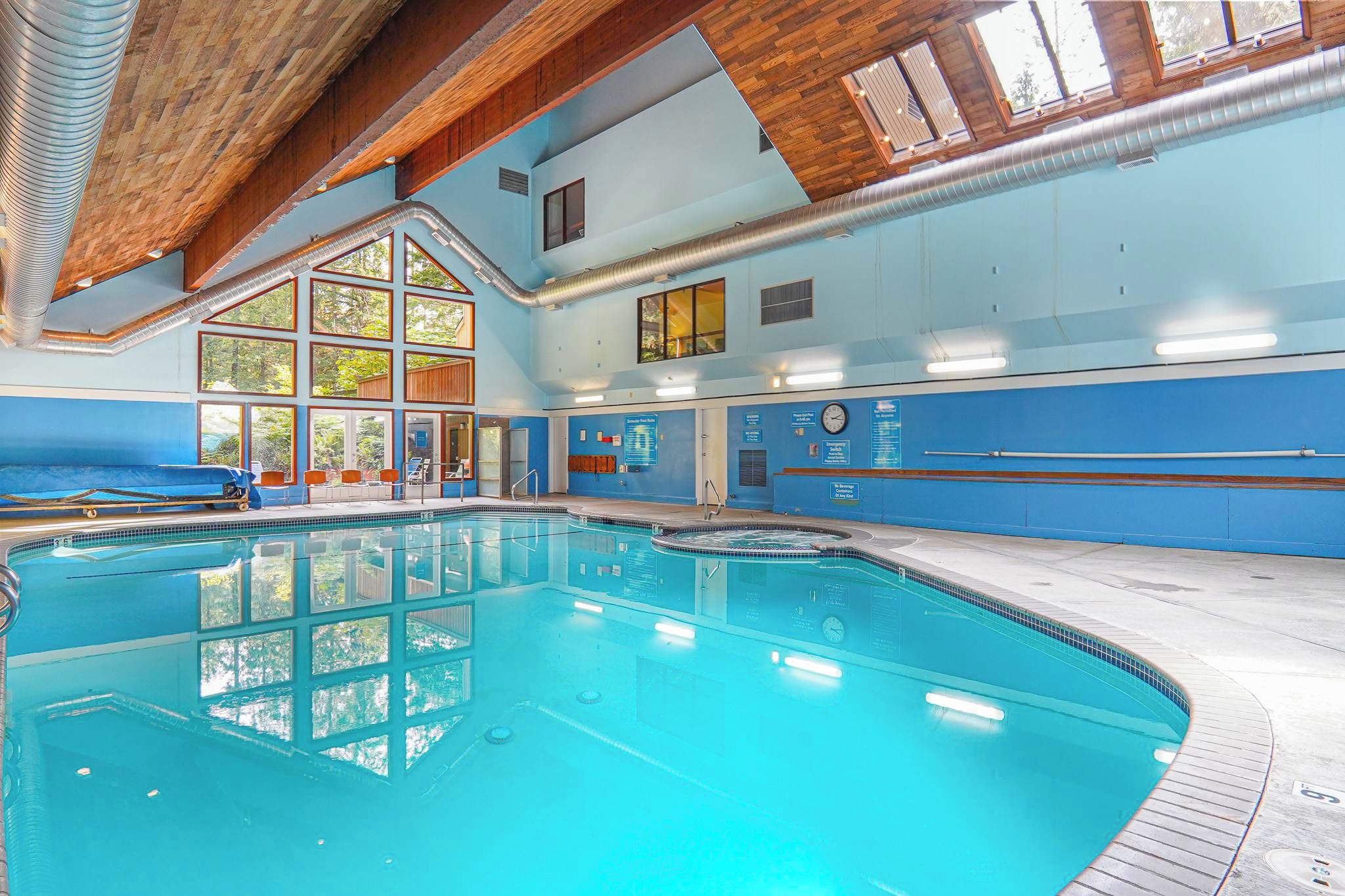 An indoor swimming pool with clear blue water, wooden ceiling beams, and large windows allowing natural light.
