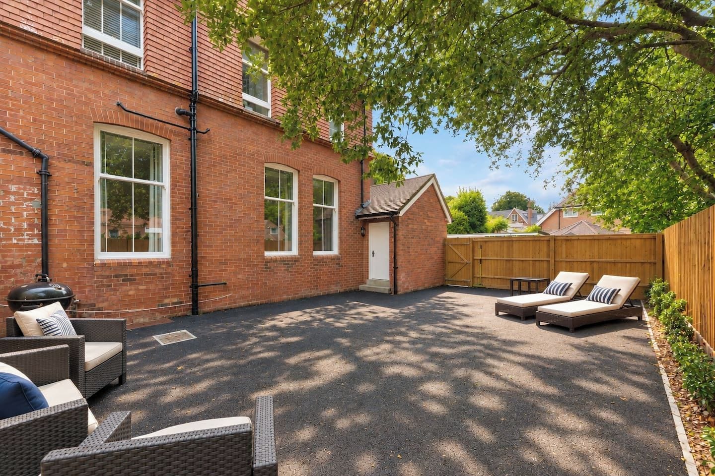 A backyard with a brick house, a wooden fence, and outdoor furniture including two lounge chairs and a wicker table.