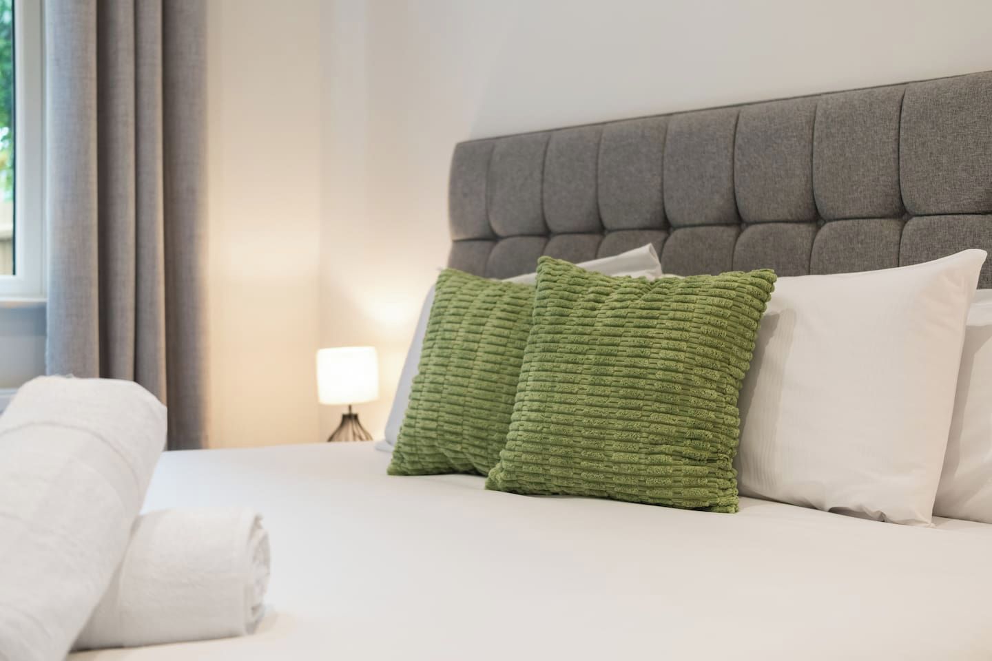 Una cama bien tendida con sábanas blancas y dos almohadas, una verde y otra blanca, en una habitación con cabecera tapizada en gris.