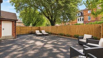 A backyard with a paved area, two lounge chairs, a table, and a wooden fence.