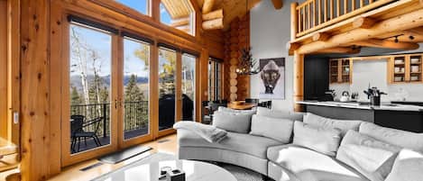 A modern log cabin interior with a large curved sofa, a dining area, and a view through large windows.
