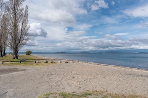 Ein Kiesstrand mit einer Holzbank, Bäumen und Blick auf das Wasser und entfernte Berge.