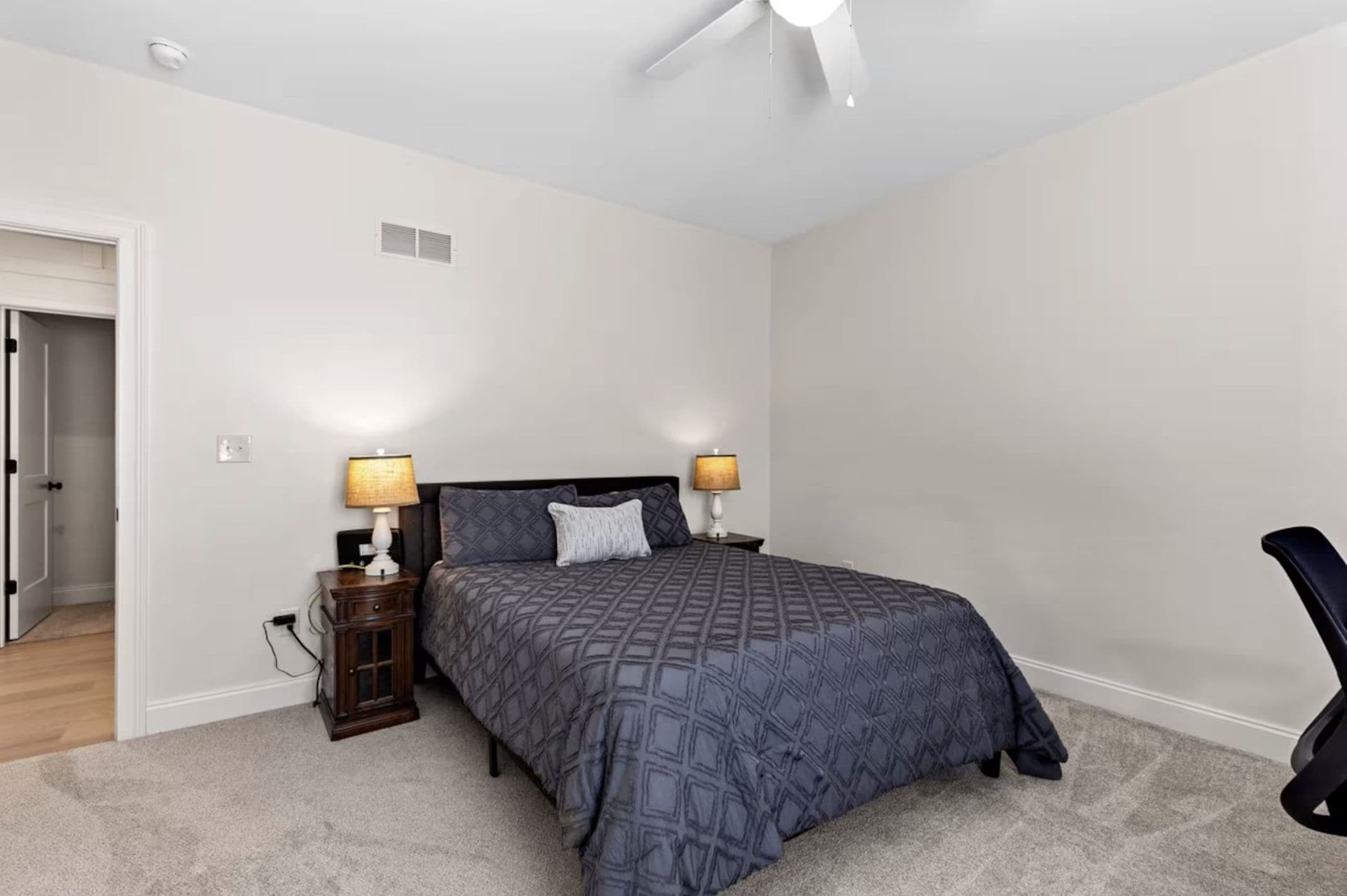 A bedroom with a bed, two bedside tables with lamps, a ceiling fan, and a door leading to another room.