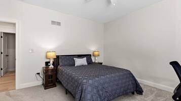 A bedroom with a bed, two bedside tables with lamps, a ceiling fan, and a door leading to another room.