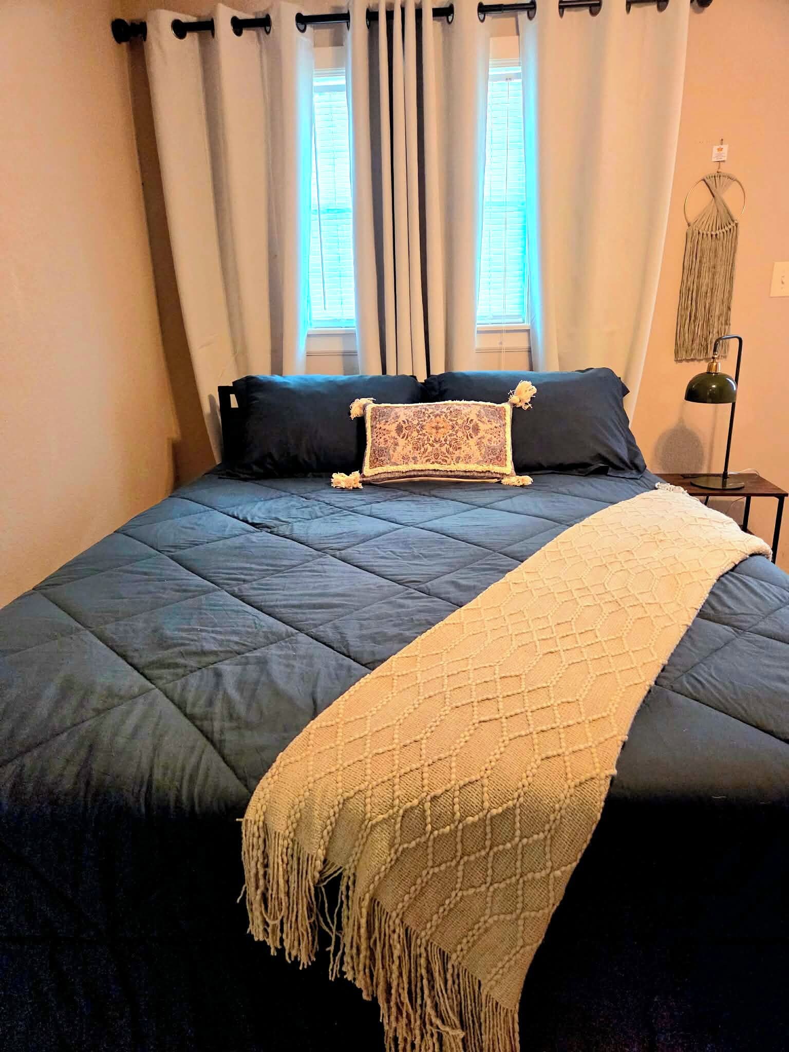 A bed with a dark blue quilt, two pillows, a decorative throw, and a wall-mounted lamp.